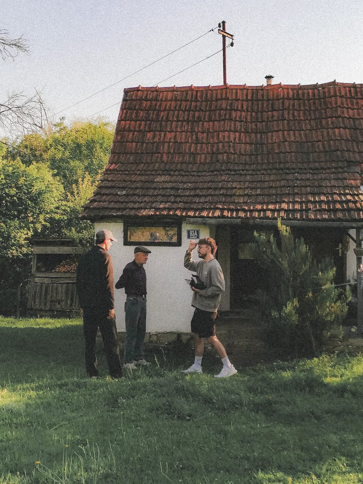 Luka Jovičić directing two actors in front of a village house, holding the director’s monitor